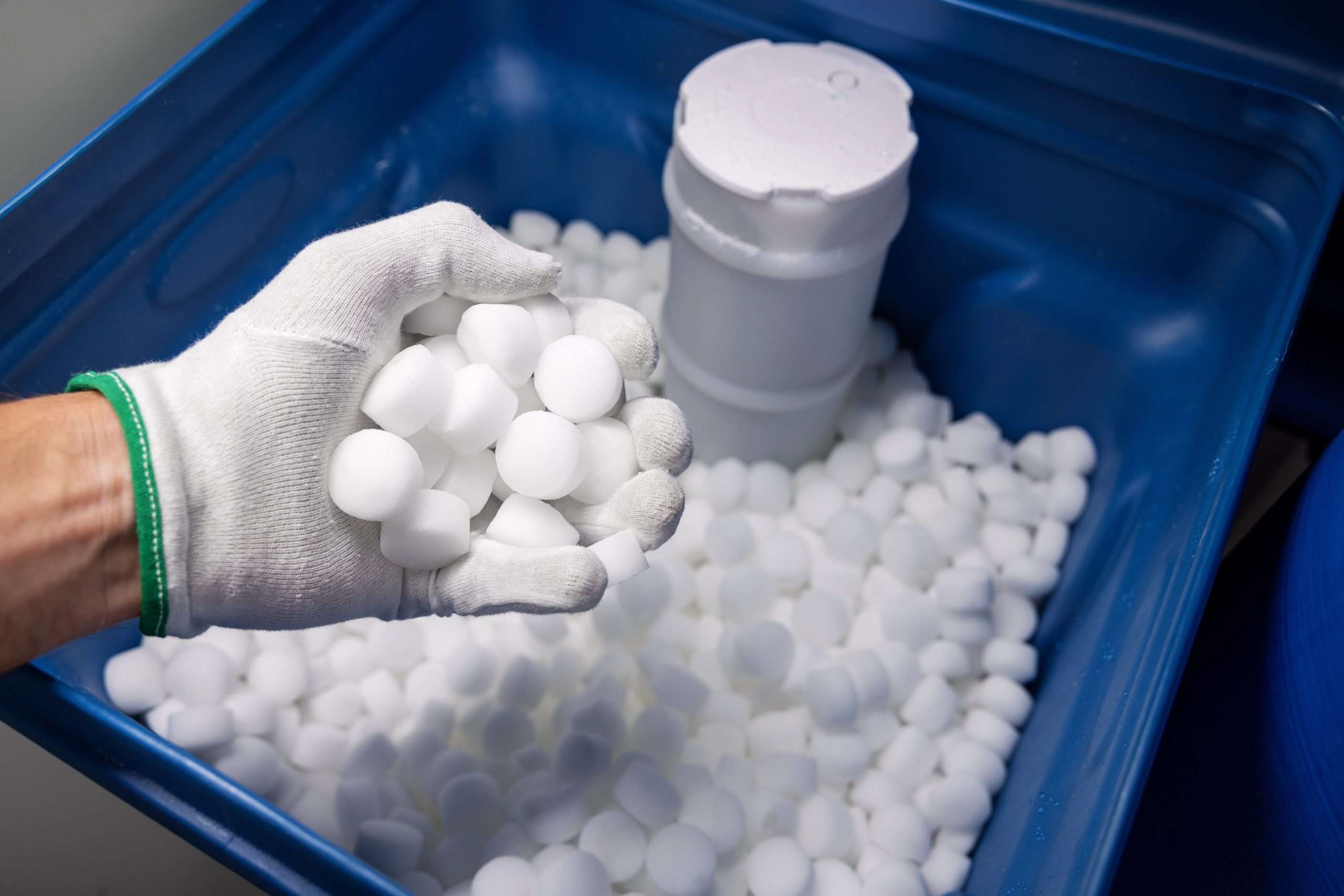 A white gloved hand holding a handful of water softener tablets over a bucket full of water softener tablets.